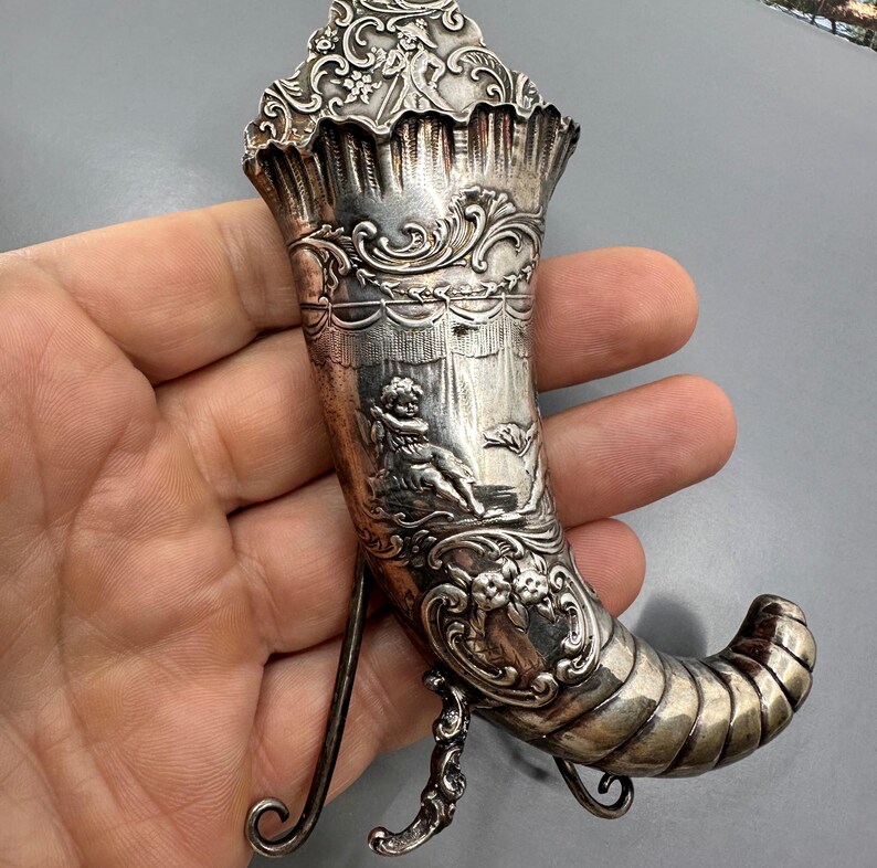 May include: A silver, ornate, decorative horn with a stand. The horn is decorated with intricate floral and cherub designs. The horn is approximately 15 cm tall.