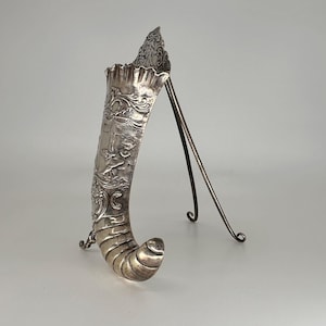 May include: A silver, ornate, horn-shaped vase with a stand. The vase is decorated with a floral design and a cherub.