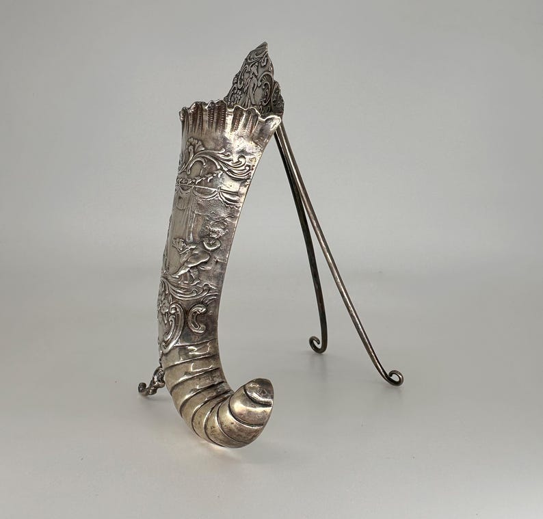 May include: A silver, ornate, horn-shaped vase with a stand. The vase is decorated with a floral design and a cherub.