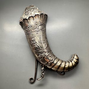 May include: A silver, ornate, decorative horn with a stand. The horn is decorated with a raised design of cherubs and floral motifs.