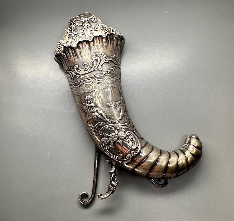 May include: A silver, ornate, decorative horn with a stand. The horn is decorated with a raised design of cherubs and floral motifs.
