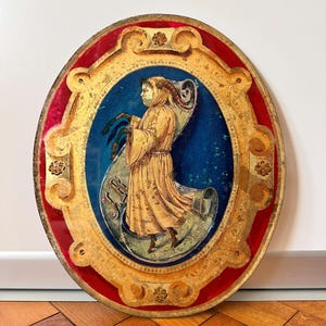 Può includere: Opera d'arte ovale vintage con cornice dorata e bordo rosso. L'opera presenta una figura in abito dorato su sfondo blu, che tiene in mano una pergamena. La cornice presenta dettagli ornati.