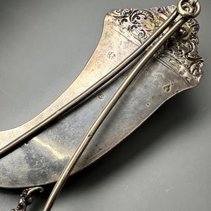 May include: A silver, crescent-shaped serving tray with a decorative handle and two attached serving spoons. The tray has an ornate design and is made of polished metal.