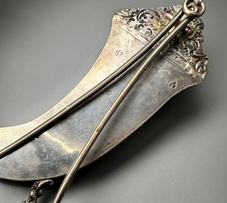 May include: A silver, crescent-shaped serving tray with a decorative handle and two attached serving spoons. The tray has an ornate design and is made of polished metal.