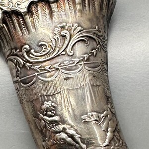 May include: A silver horn-shaped cup with intricate embossed designs. The cup features cherubs and a draped fabric design.