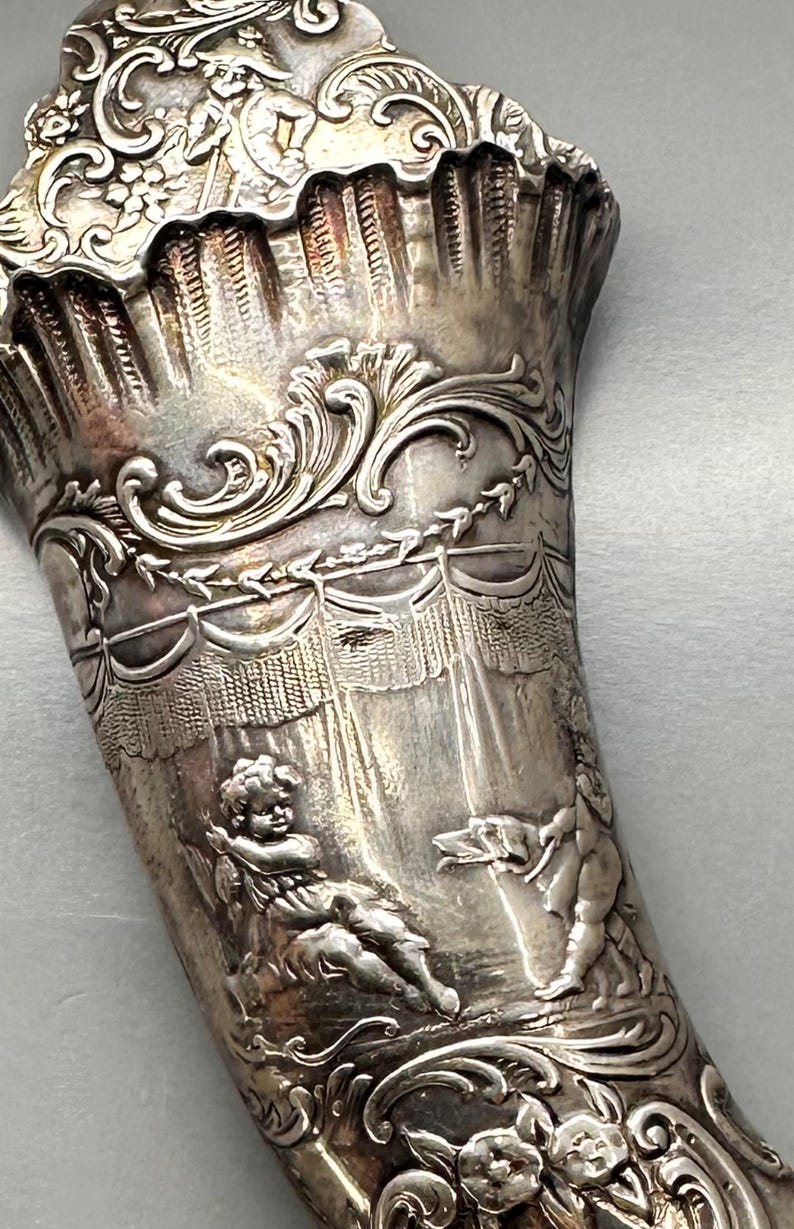 May include: A silver horn-shaped cup with intricate embossed designs. The cup features cherubs and a draped fabric design.