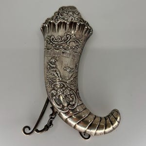 May include: A silver horn-shaped container with intricate engravings. The horn is decorated with a scene of a cherub and a dog. The container has a hinged lid and a chain attached to the bottom.