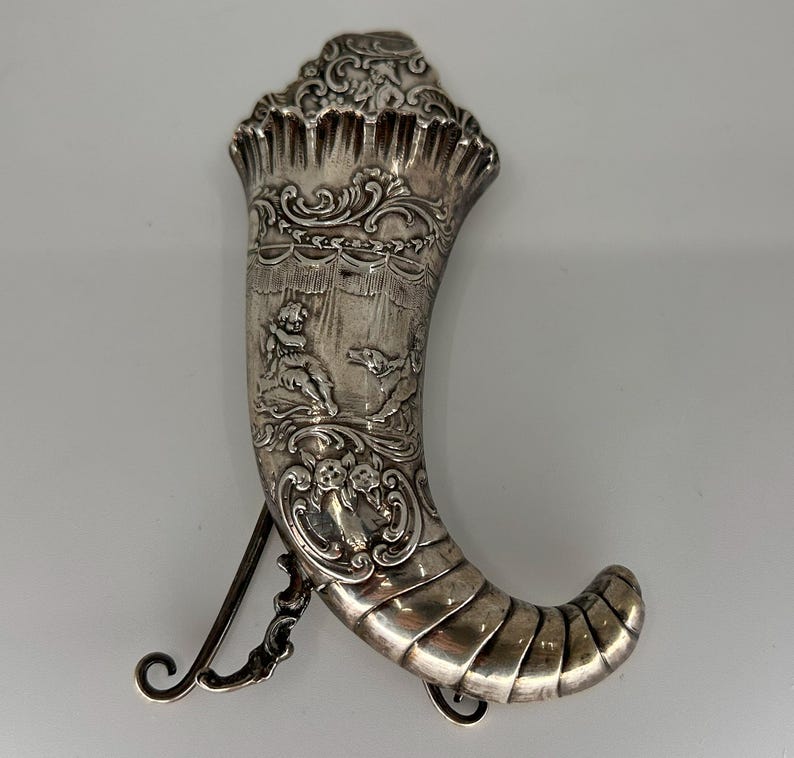 May include: A silver horn-shaped container with intricate engravings. The horn is decorated with a scene of a cherub and a dog. The container has a hinged lid and a chain attached to the bottom.