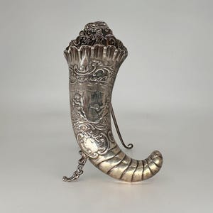 May include: A silver, ornate, decorative horn with a stand. The horn is decorated with a scene of two figures and intricate scrollwork.