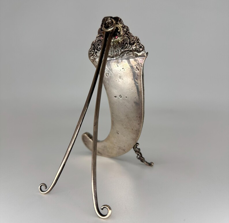 May include: Silver picture frame with a decorative top and a stand. The frame is shaped like a horn and has a silver chain hanging from the bottom.