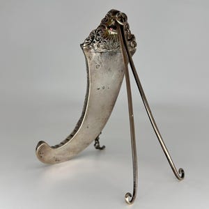 May include: A silver picture frame with an ornate design and a stand. The frame is shaped like a crescent moon and has a small red stone at the top.