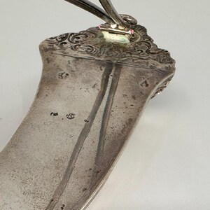 May include: A silver serving spoon with an ornate handle and a curved bowl. The spoon has a maker's mark on the back of the handle.