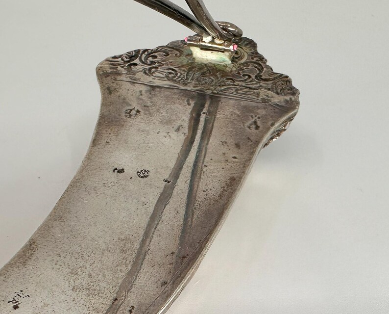 May include: A silver serving spoon with an ornate handle and a curved bowl. The spoon has a maker's mark on the back of the handle.