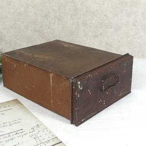 Op de afbeelding: Een rechthoekige, antieke houten lade met een metalen handvat. De lade is een mix van bruin en donkerbruin, met zichtbare slijtage. Een vintage document ligt op de voorgrond, met groen op de achtergrond.