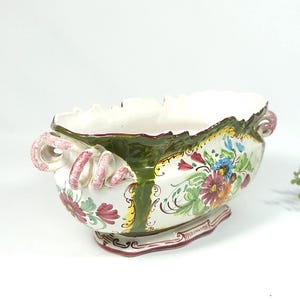 Può includere: Fioriera ovale in ceramica con bordo smerlato e design floreale. La fioriera presenta un interno bianco, dettagli verdi e gialli e manici rosa. L'esterno è decorato con fiori e foglie colorate.