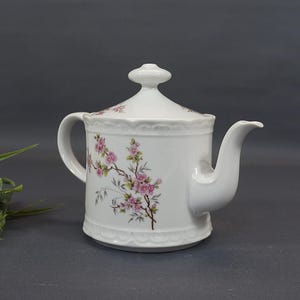 Könnte beinhalten: Weiße Keramik-Teekanne mit Deckel, Henkel und Ausguss. Die Teekanne ist mit rosa Blütenzweigen verziert. Der Boden und der Deckelrand haben ein gewelltes Design. Die Teekanne steht vor einem grauen Hintergrund.