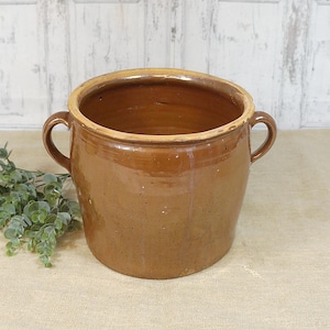 Puede incluir: Una olla de cerámica marrón con dos asas. La olla tiene una abertura ancha y un borde ligeramente más claro. El exterior tiene un acabado brillante. Una ramita de follaje verde descansa junto a la olla.