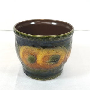 Può includere: Un piccolo vaso in ceramica verde con un bordo marrone e un motivo floreale in tonalità di giallo, arancione e verde.