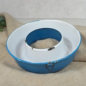 Op de afbeelding: Een ronde, blauw met witte geëmailleerde metalen tulbandvorm met een centrale buis. De buitenkant is blauw, terwijl de binnenkant wit is. De rand en de centrale buis vertonen gebruikssporen met kleine plekken waar het emaille is afgebladderd.