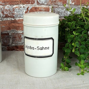 Puede incluir: Dos tarros de cerámica blanca con etiquetas negras. El tarro de la derecha tiene una etiqueta que dice "Jojoba-Sahne".