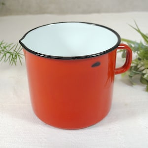 Könnte beinhalten: Eine rote Emaille-Tasse mit weißem Innenraum und schwarzem Rand. Die Tasse hat einen Henkel und steht auf einer weißen Oberfläche.