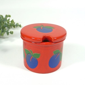 Può includere: Un barattolo di ceramica rosso con coperchio abbinato. Il barattolo e il coperchio sono decorati con disegni di prugne blu e verdi. Il coperchio ha una piccola tacca per un cucchiaio. Il barattolo è probabilmente per conservare conserve o condimenti.
