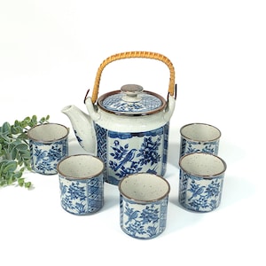 Könnte beinhalten: Keramik-Teeservice mit einer Teekanne und sechs passenden Tassen. Die Teekanne hat einen geflochtenen Griff und ist mit blauen Blumen- und Vogelmustern auf weißem Hintergrund verziert. Das Set hat eine gesprenkelte, strukturierte Oberfläche.