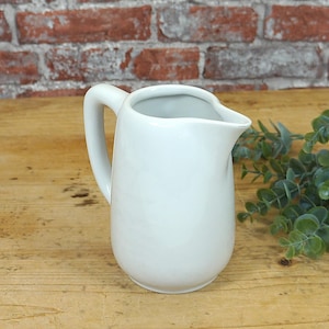 Könnte beinhalten: Ein weißer Keramik-Krug mit Henkel. Der Krug steht auf einer Holzoberfläche.