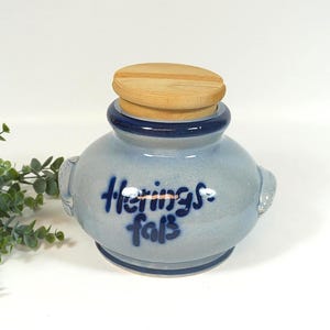 Può includere: Un barattolo di ceramica azzurra con coperchio in legno. Il barattolo ha un bordo e manici blu scuro. Il testo "Herings faß" è scritto in blu scuro sul davanti del barattolo. Un rametto di foglie verdi è sullo sfondo.