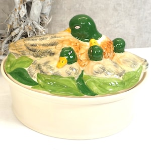 Op de afbeelding: Een keramische serveerschaal met een deksel met een handgeschilderd ontwerp van eenden. De eenden zijn geschilderd in tinten bruin, groen en geel. De schaal is wit met een groene rand.