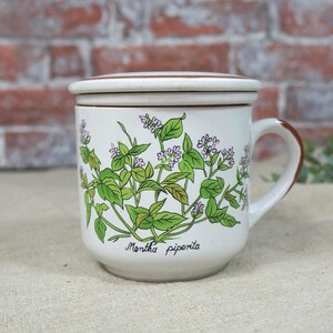Può includere: Tazza in ceramica bianca con coperchio, con un'illustrazione botanica di foglie verdi e fiori viola. La tazza ha un manico e un bordo marrone, con la scritta "Mentha piperita". Un'aggiunta affascinante a qualsiasi cucina.