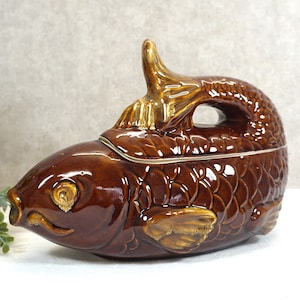 Könnte beinhalten: Eine braune, fischförmige Servierplatte aus Keramik mit Deckel. Der Deckel hat einen goldenen Griff und der Fisch hat goldene Akzente an den Flossen und Augen.