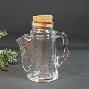 Op de afbeelding: Een heldere glazen kan met een getextureerd oppervlak, een handvat en een tuit. De kan is afgedekt met een kurken stopper. De achtergrond is donkergrijs en er is groen links van de kan.