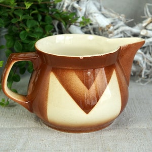 Op de afbeelding: Een vintage bruin en crèmekleurige keramische kan met een geometrisch ontwerp. De kan heeft een handvat en een tuit, en de binnenkant is crèmekleurig. De buitenkant heeft een bruin en crèmekleurig chevronpatroon. Een charmante toevoeging aan elke keuken.