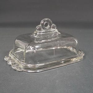 Op de afbeelding: Een glazen botervloot van helder glas met een rechthoekige bodem en een bijpassend deksel. Het deksel heeft een centrale boogvormige handgreep met een ronde opening aan de bovenkant. De randen van de bodem en het deksel hebben een parelrand.