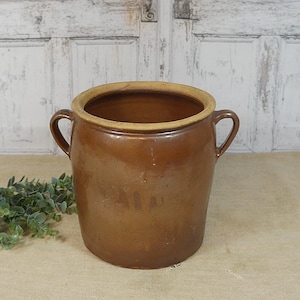 Puede incluir: Un tarro de cerámica marrón con dos asas y un borde de color beige claro. La olla antigua es cilíndrica y se asienta sobre una superficie neutra. Una ramita de follaje verde está a la izquierda del tarro.