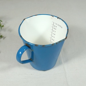 Può includere: Tazza dosatrice in smalto blu con interno bianco e manico. La tazza ha una capacità di 1,0 litro, con misurazioni a incrementi di 0,1 litro. La tazza ha un aspetto leggermente usurato.