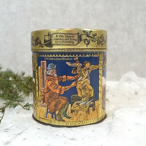 Könnte beinhalten: Eine vintage Blechdose mit blauem Hintergrund und goldenen Verzierungen. Die Dose zeigt eine Szene mit zwei Figuren, eine hält einen Hammer und die andere ein Stück Holz. Der Text auf der Dose lautet "E-Otto Schmidt Lebkuchen und Honig 8500 Nürnberg" und "Der Stoff in feiner Werkstatt".