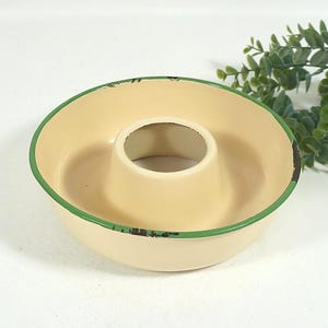 Op de afbeelding: Een ronde, crèmekleurige emaille bakvorm met een centrale buis en een groene rand. De rand vertoont zichtbare donkere, versleten plekken langs de rand, waardoor het metaal eronder zichtbaar is. De binnenkant is glad en licht van kleur.