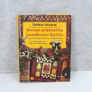 Op de afbeelding: Een boek getiteld "Debbie Mumm: Meine schönsten Landhaus-Quilts" met een gele kaft en kleurrijke illustraties van patchwork dekens, boerderijdieren en bloemen. Het boek bevat meer dan 70 eenvoudige patchwork- en decoratie-ideeën met professionele tips voor snel nawerken.