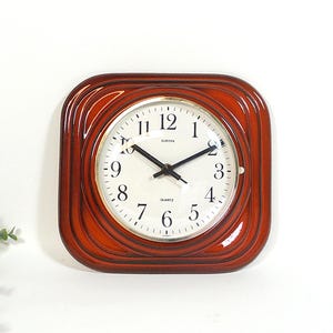 Könnte beinhalten: Quadratische Wanduhr mit Holzrahmen in Rotbraun. Das Zifferblatt ist hell mit schwarzen Ziffern und Zeigern. Das Wort "EUROPA" befindet sich über der Mitte. Klassisches Design.