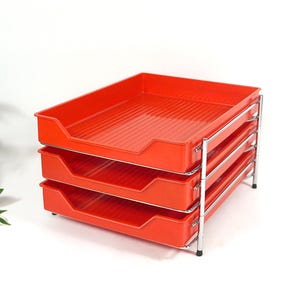 Könnte beinhalten: Ein dreistufiger Schreibtisch-Organizer mit leuchtend orangefarbenen Kunststoffschalen, die eine gerippte Innenfläche aufweisen. Die Schalen sind vertikal gestapelt und werden von einem glänzenden Chrom-Metallrahmen mit vier dünnen Beinen gehalten.