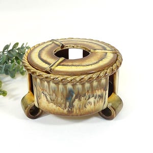 Puede incluir: Un cenicero de cerámica con un diseño circular y un borde en forma de cuerda. El cenicero tiene una combinación de colores marrón y amarillo con un acabado brillante. Cuenta con una abertura central y descansa sobre tres patas curvas.