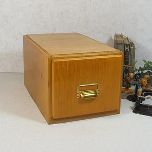 Puede incluir: Un archivador de madera marrón claro con un tirador de latón. El mueble tiene forma rectangular y un diseño sencillo. Es una pieza vintage, perfecta para organizar documentos o añadir un toque rústico a una oficina en casa. Mide aproximadamente 30 cm de alto.