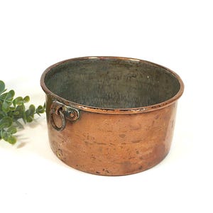 Puede incluir: Una olla redonda de cobre antiguo con un interior deslustrado y un pequeño asa en el lateral. La olla tiene un aspecto rústico y envejecido, lo que sugiere que es vintage. La olla está sobre un fondo blanco con una ramita de follaje verde.