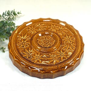 Könnte beinhalten: Ein runder, brauner Keramik-Topfuntersetzer mit floralem Design. Der Topfuntersetzer hat ein erhabenes, geprägtes Muster aus Blumen und Blättern am Rand und in der Mitte. Der Topfuntersetzer hat eine warme, erdige braune Farbe und eine glänzende Oberfläche. Der Topfuntersetzer hat einen Durchmesser von etwa 20 cm.