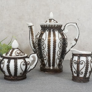 Op de afbeelding: Een wit porseleinen theeservies met ingewikkelde gouden filigraanontwerpen. De set bevat een theepot, een roomkan en een suikerpot.