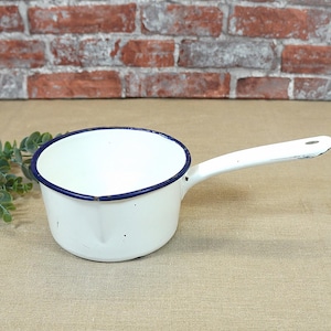 Op de afbeelding: Een witte emaille pan met een blauwe rand en een handvat. De pan staat op een beige stoffen oppervlak.