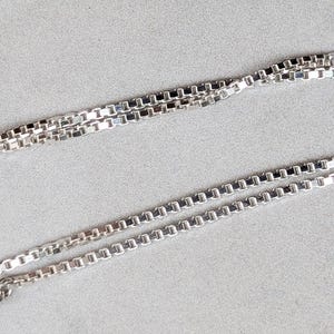 Peut inclure: Un collier en chaîne argent avec un design de chaîne à maillons carrés. La chaîne est présentée en deux couches.