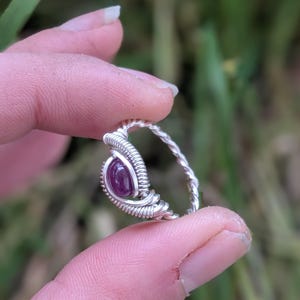 May include: A silver wire wrapped ring with a purple gemstone. The ring is on a finger.